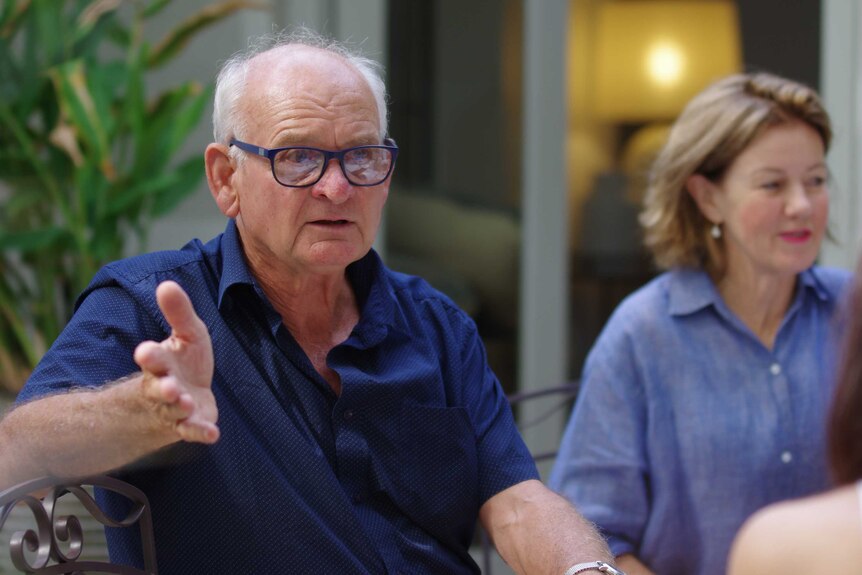 Man sits at outdoor table, woman in the background, talking with arm waving