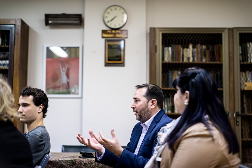 Sacha D'Souza gestures with hands while speaking in a white fluorescent lit meeting room with caged bookcases.