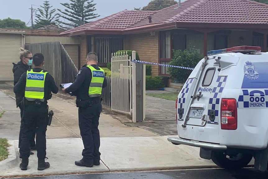 Two uniformed police officers speak to a masked detective outside a suburban house.