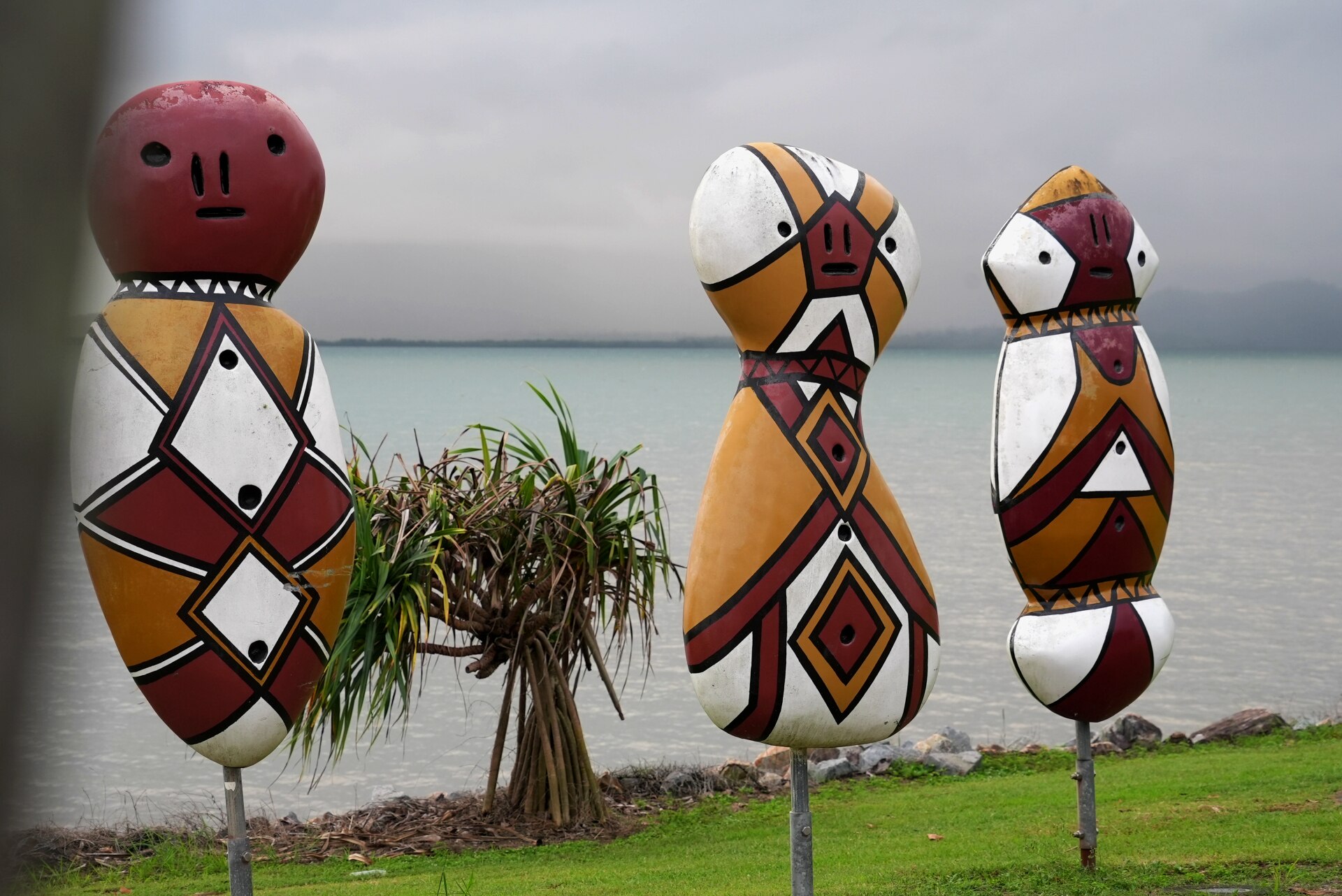 Three large body and head-shaped painted sculptures on metal poles, standing on grass with sea and islands behind.