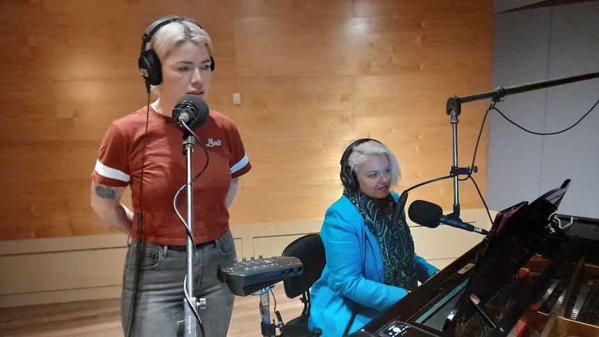 a woman stands and sings into a microphone, while another woman is seated playing a keyboard