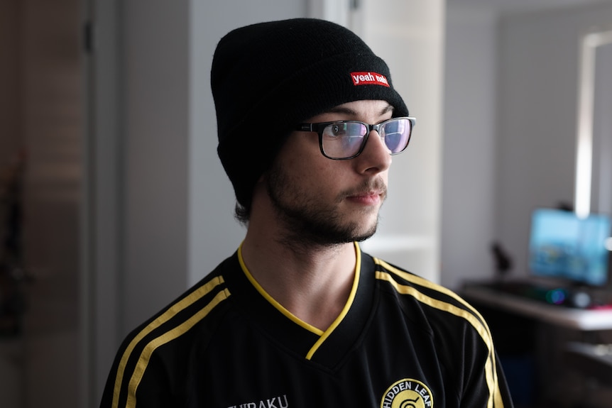 A portrait of a teenager, standing indoors, wearing a beanie and glasses. He is looking to the side with a neutral expression.