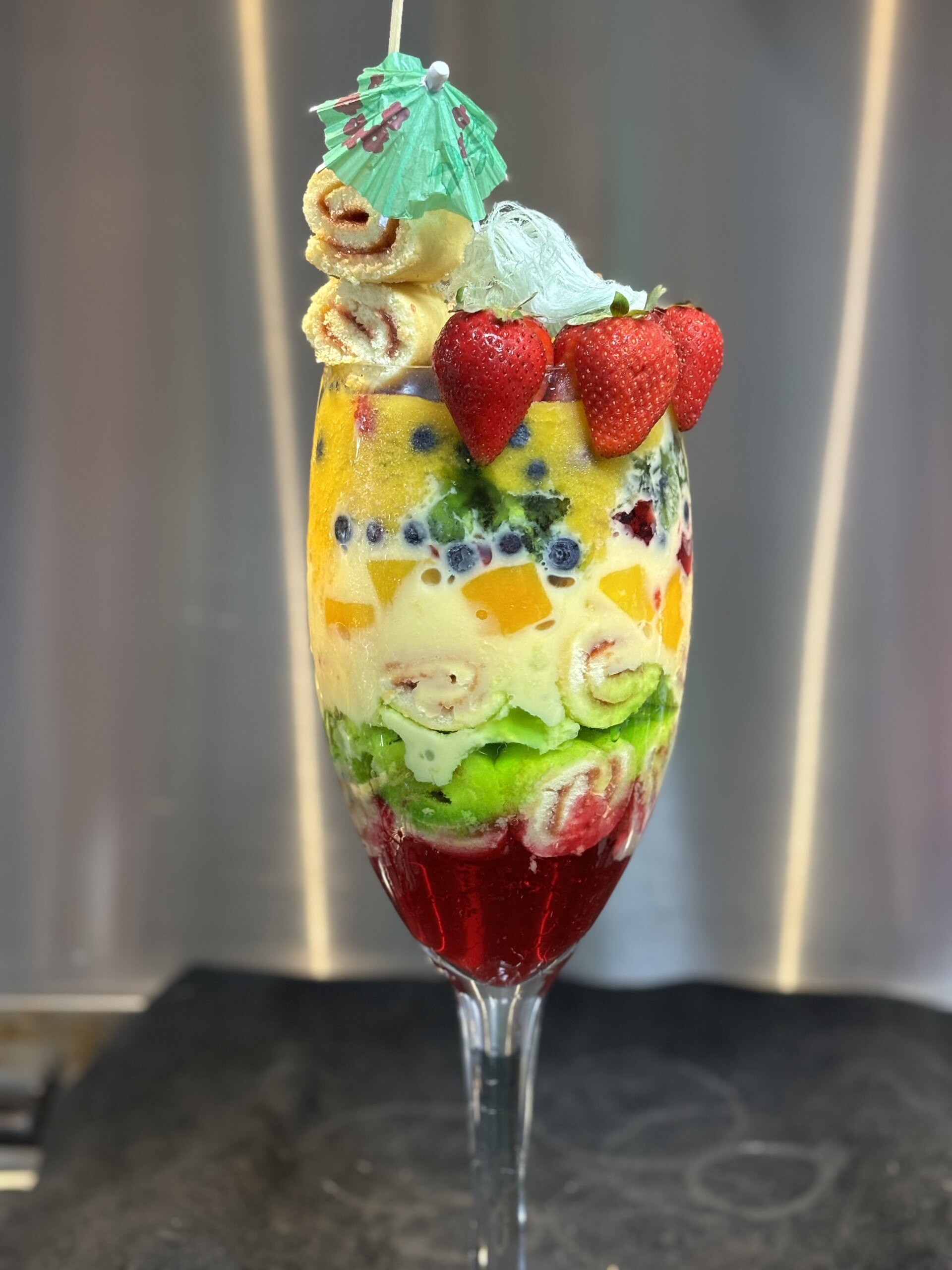 A wine glass filled with trifle layers of jelly, jam roll slices, custard, berries and mango puree, served with blue fairy floss