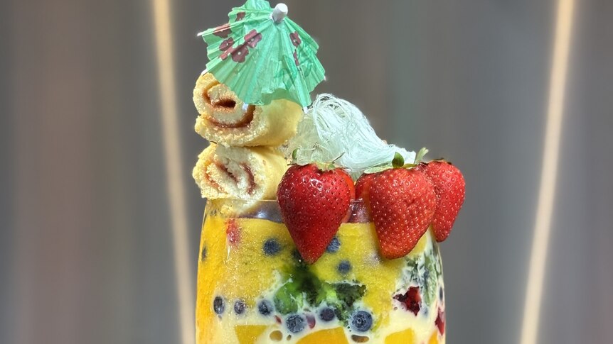 A wine glass filled with trifle layers of jelly, jam roll slices, custard, berries and mango puree, served with blue fairy floss