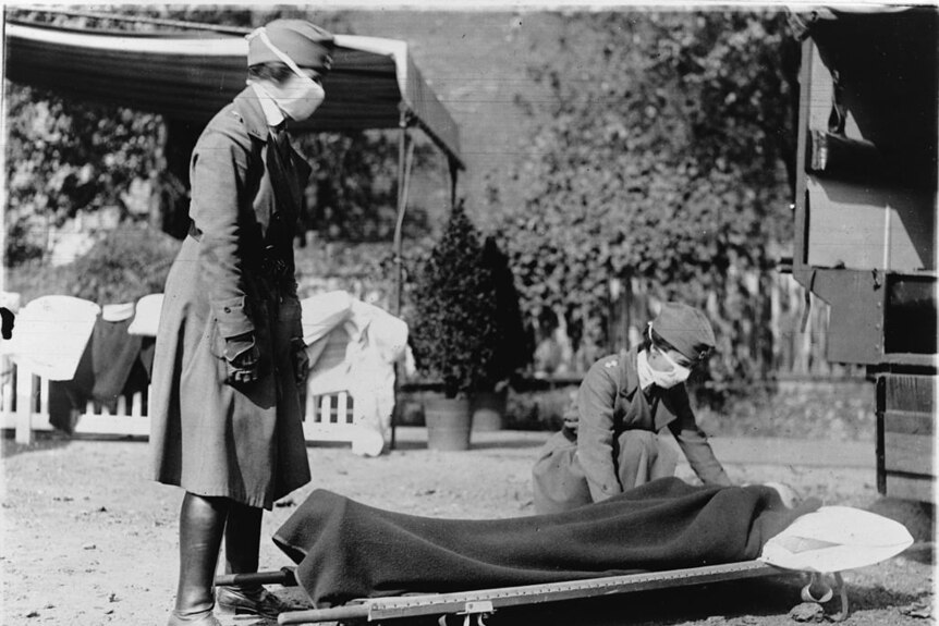 A black and white photo of women wearing nurses uniforms and face masks tending to a stretcher.