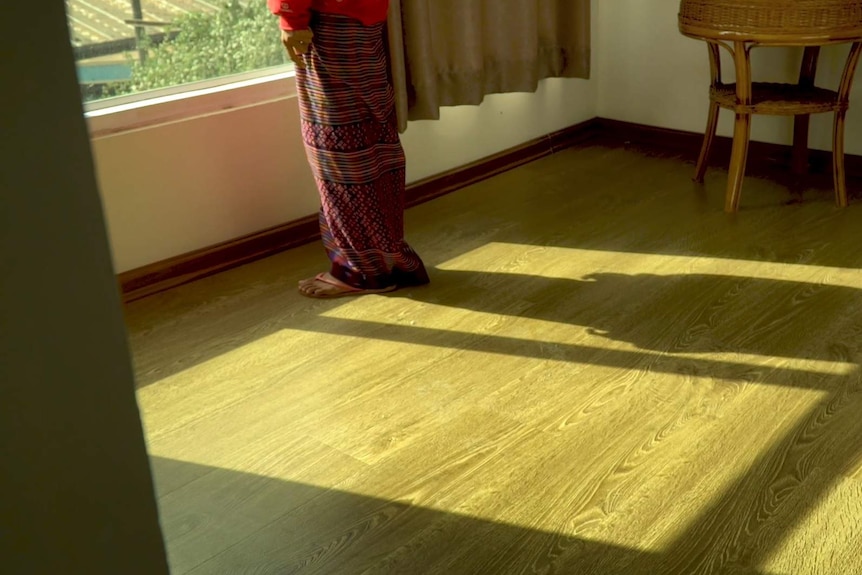 A trafficked woman from Myanmar's Kachin State looks out window. She is wearing a long purple patterned skirt and a red jumper.