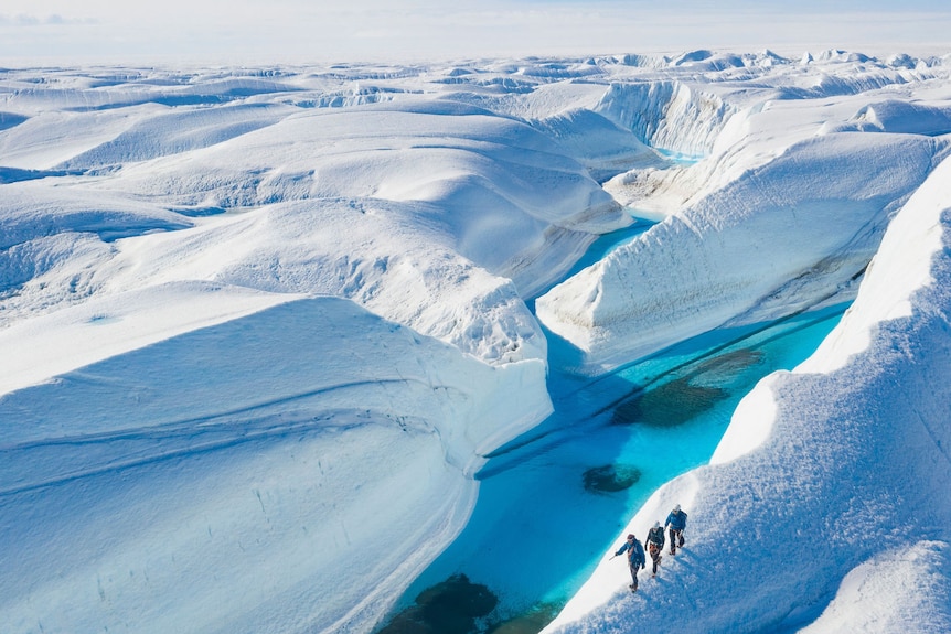 Three hikers walk along a ridge of snow past a clear blue lake with endless snow stretching in to the background.