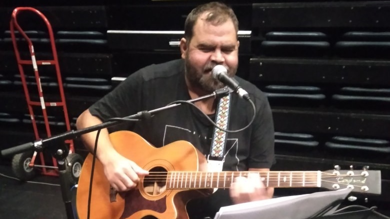 Indigenous man sitting playing guitar and singing into microphone with music sheets on stand