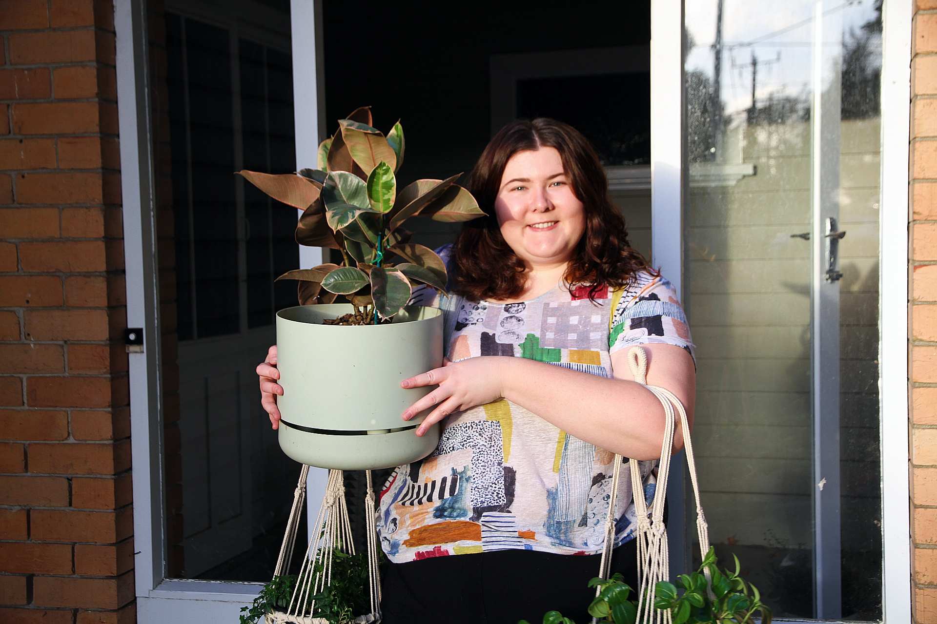 A woman juggles three plants in the doorway of a brick house.