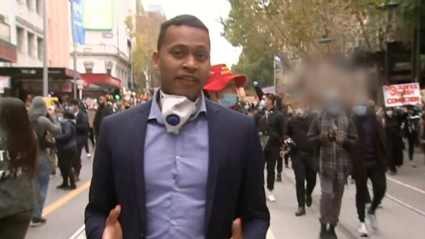 Clure with face mask around neck walking in front of protestors.