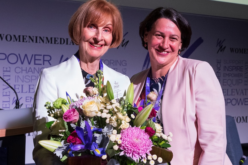 Carol jones stands with a large bunch of pink and purple flowers next to woman in a pink blazer