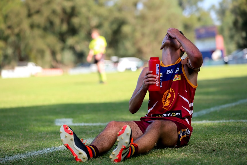 Young footballer leans back and sucks on a soy sauce sachet while holding a water bottle and sitting on football ground sideline