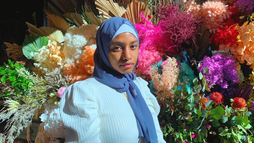 A woman wearing the hijab standing in front of a floral background.