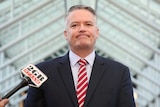 Mathias Cormann, wearing a pinstriped suit and red and white patterned tie, grimaces as reporters nudge microphones towards him.