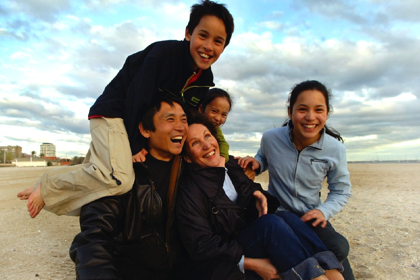 A family consisting of mum, dad, son and two daughters sit together close, all laughing, at the beach