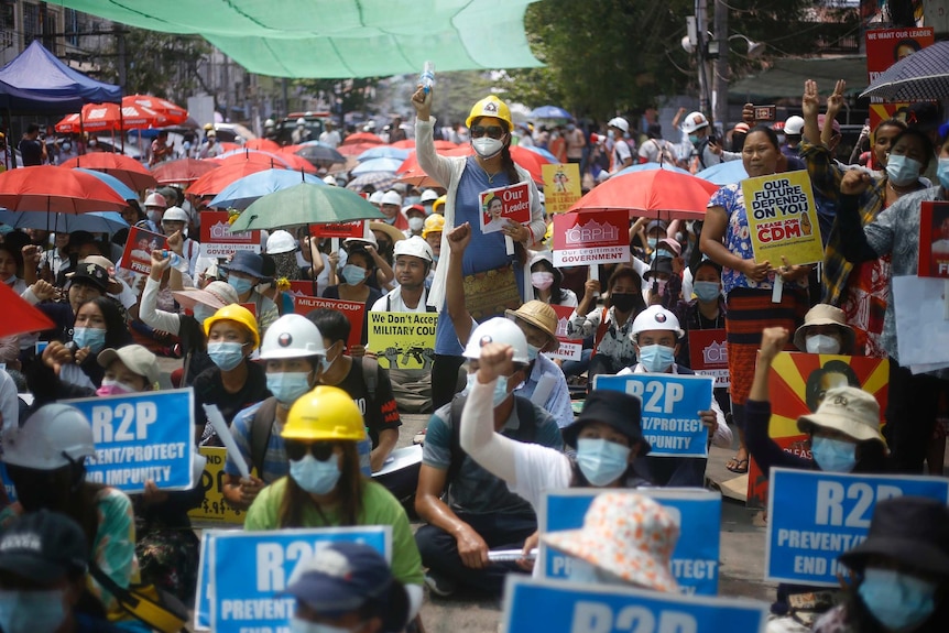A large crowd of protesters sit under umbrellas and hold signs picturing Aung San Suu Kyi.