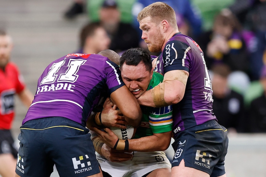 A Canberra Raiders NRL player holds the ball while being tackled by two Melbourne Storm opponents.