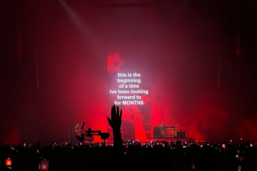 A red lit stage with big screens shows a man smiling and playing piano with white text over the top