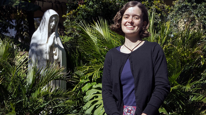 A woman stands in a garden in front of a statue of Mary