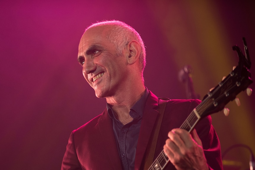 Paul Kelly smiles widely with bright lights behind him. The top of his guitar can be seen in his hands.