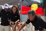 Four Indigenous people work on a yacht while an Indigenous flag flies in the background.