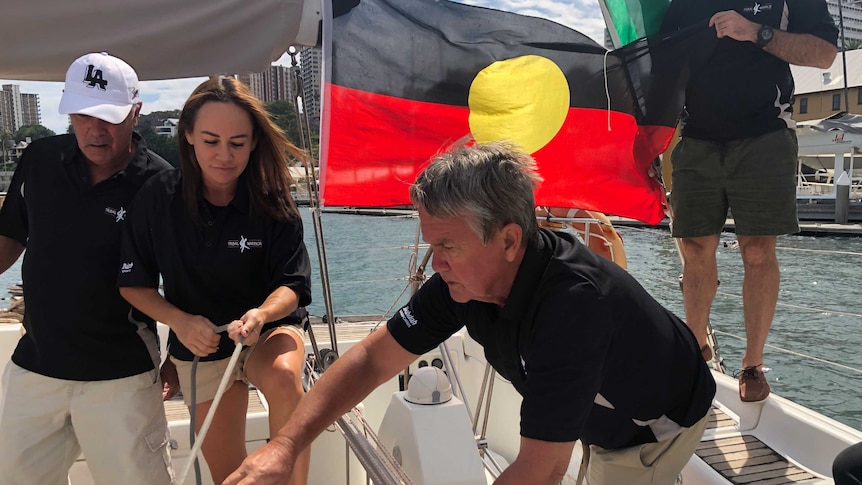Four Indigenous people work on a yacht while an Indigenous flag flies in the background.