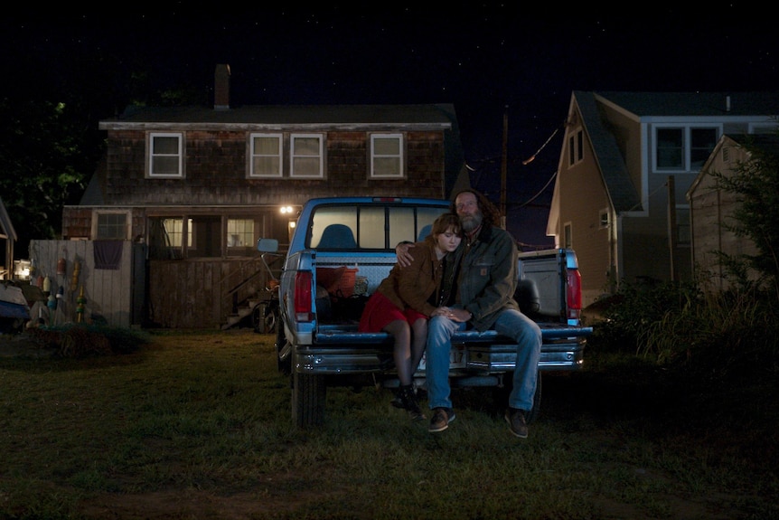 A teenage girl and her father sit together in the back of a blue truck which is parked in the backyard of a suburban house