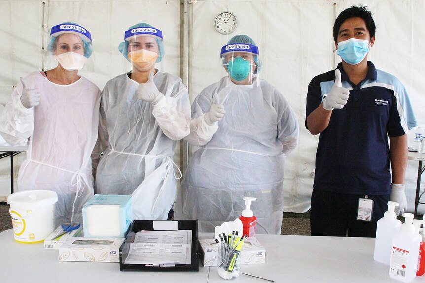 Three people in full PPE and one man in a mask standing in front of health equipment with a thumbs up.