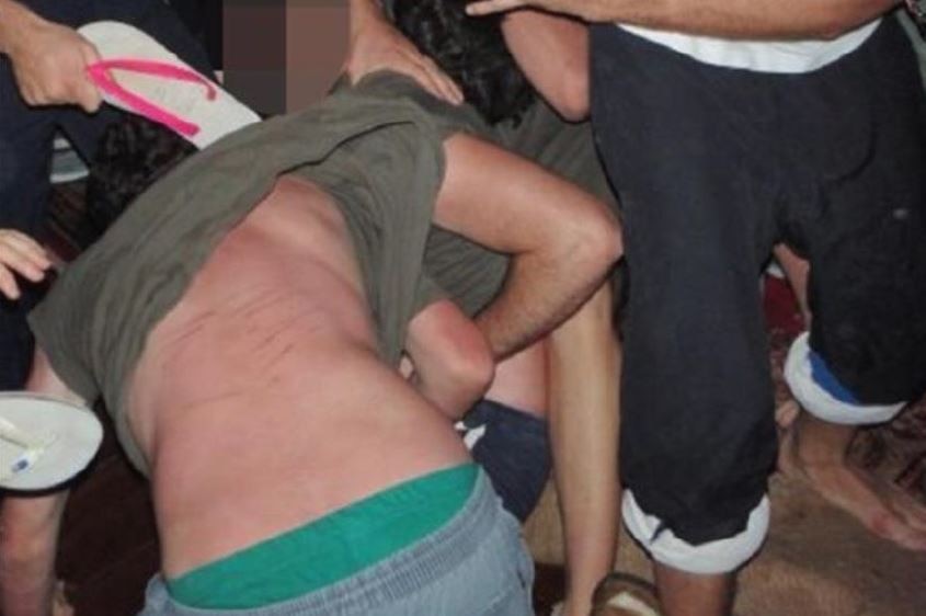 A man has red welts on his back while others stand around him holding thongs in their hands