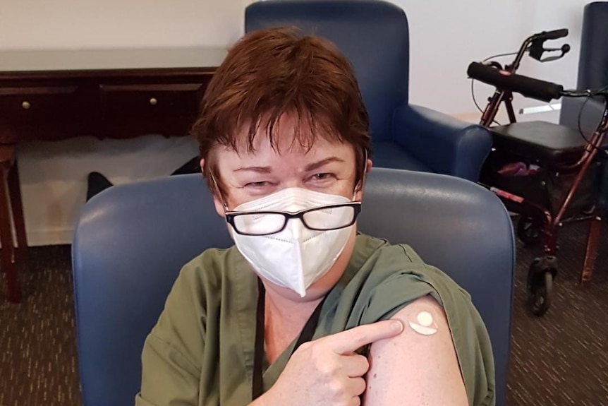 A woman wearing a mask points to a bandaid on her arm.