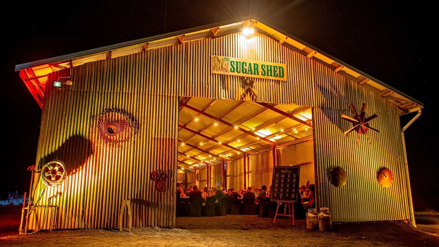 Shed with "The Sugar Shed" sign lit up at night with people seated inside