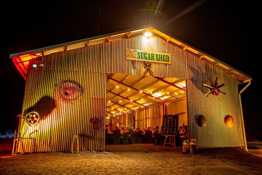 Shed with "The Sugar Shed" sign lit up at night with people seated inside