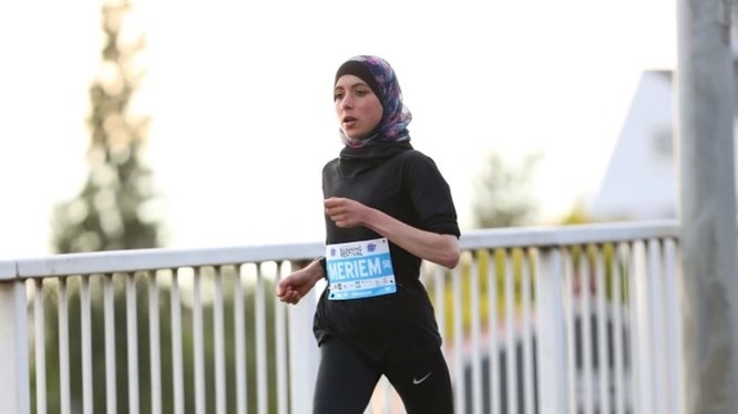 Woman wearing a hijab running in a marathon with a race number pinned to her chest that says Meriem