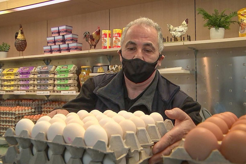 A man wearing a black face mask and black clothing puts down a pallet of eggs on a shop bench.