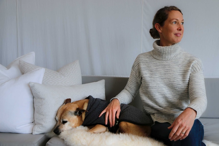 A blonde-haired woman wearing grey jump sits on a couch, patting a dog that's staring at the ground.