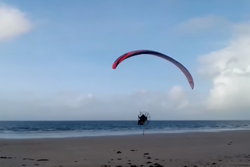 A powered paraglider, complete with parachute, carries a man up from the beach above small blue waves