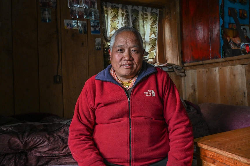 A man in a red fleece sits in a room with wood panelled walls and a bed behind