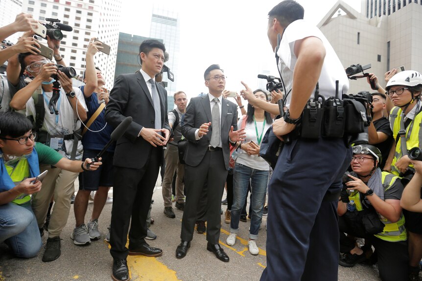 Two middle-aged men in suits stand on a  street surrounded by media as they speak with a police officer who is pointing at them.