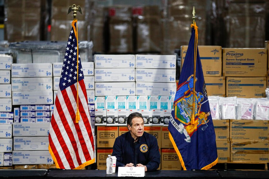 Cuomo sits in a uniform next to two large flags in front of large boxes of supplies.