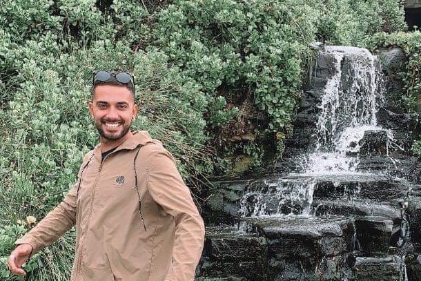 Man with brown jacket poses in front of fountain  