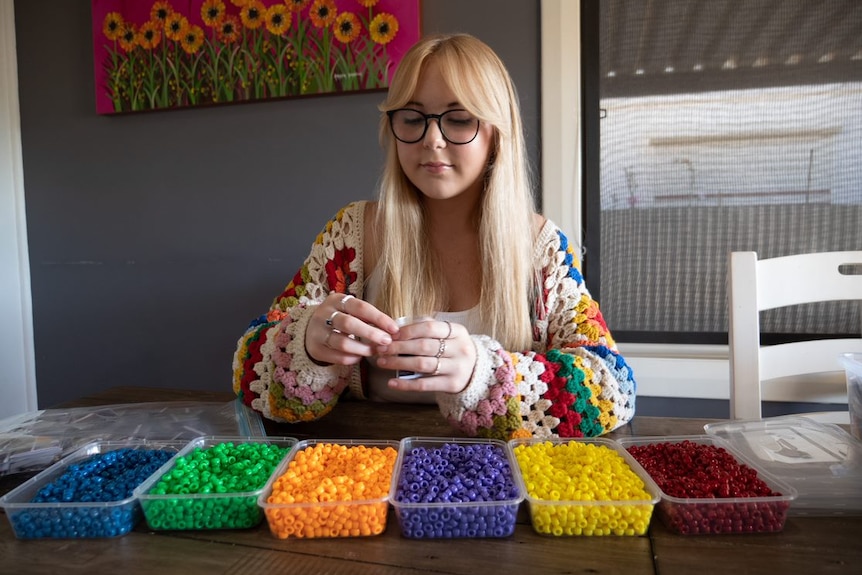 A young person with long blonde hair sits at a table with a row of containers holding different colour beads in front of her.