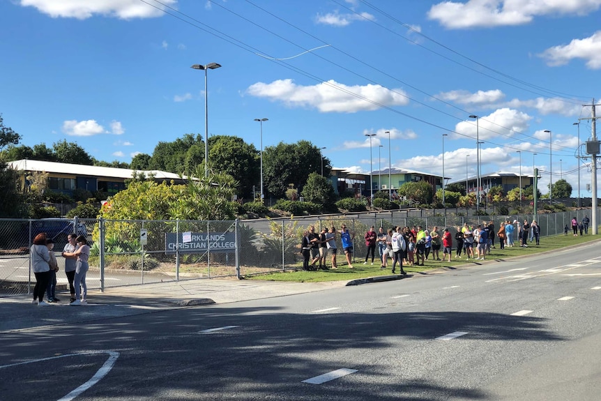 A queue of people wait outside a school fence.