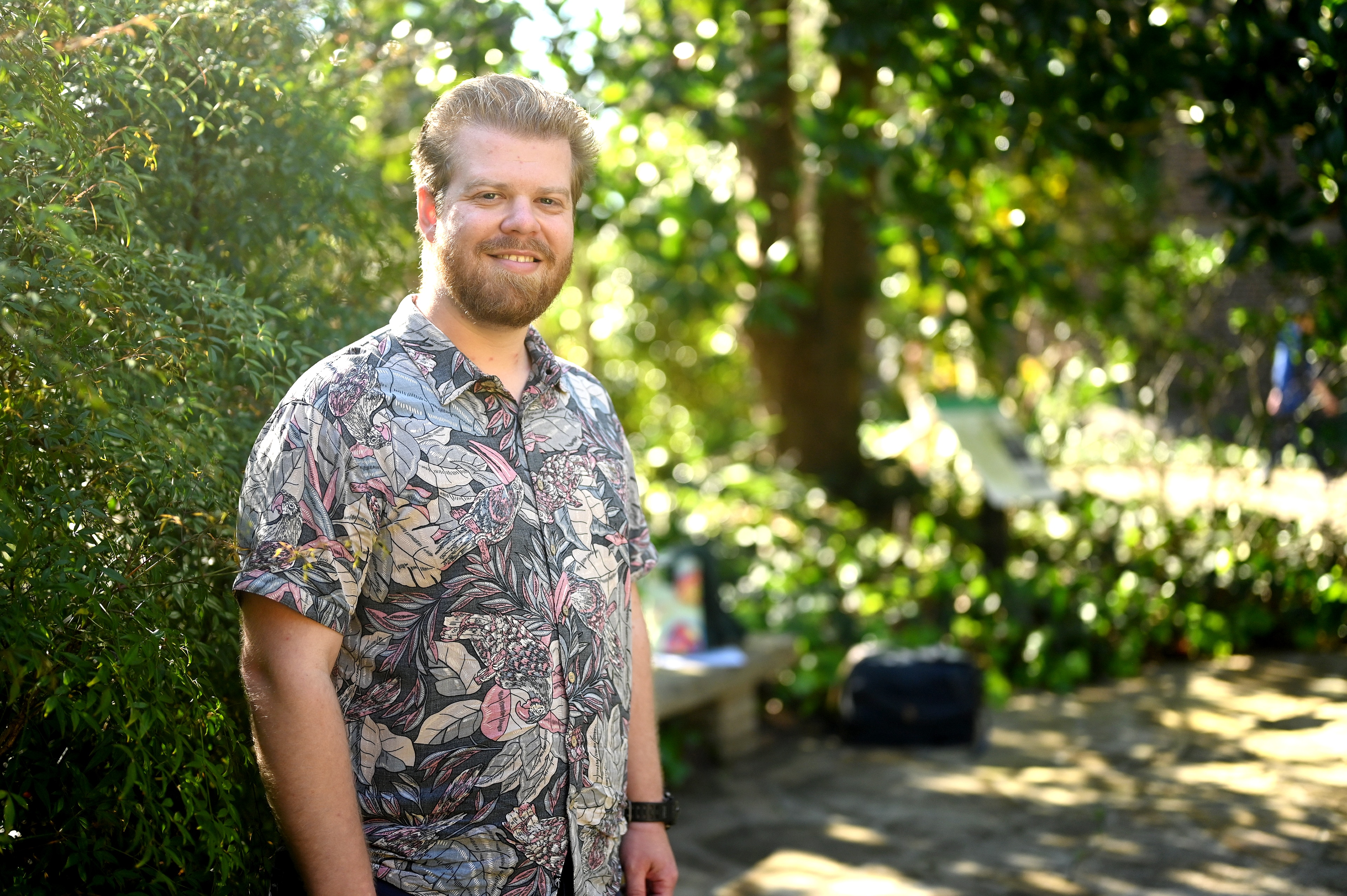 A white man with short blonde hair and bear in a grey floral shirt standing in green garden.