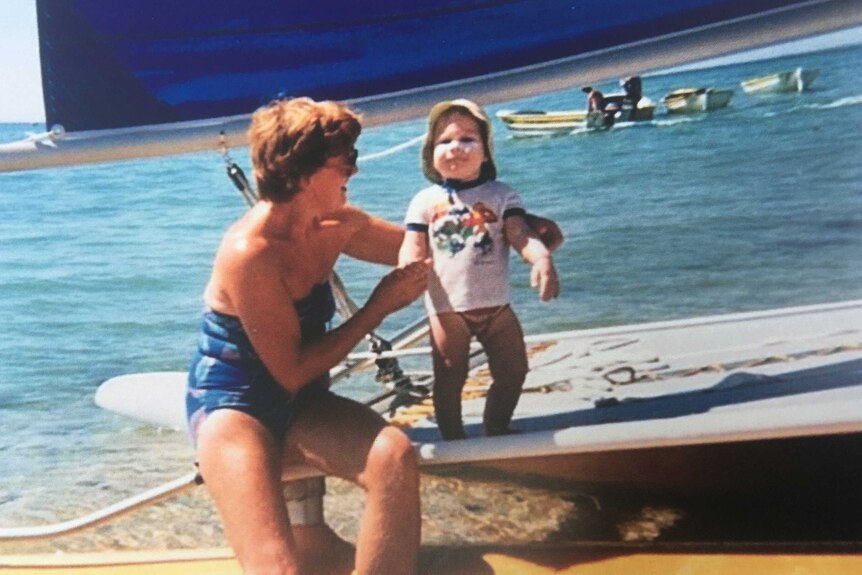 Nancy at the beach on the shore holding one of her grandchildren on a small catamaran.
