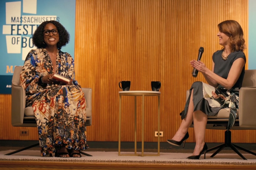 Issa Rae as Sintara Golden on stage seated, holding a book, wearing reading glasses, a woman with a mic next to her 