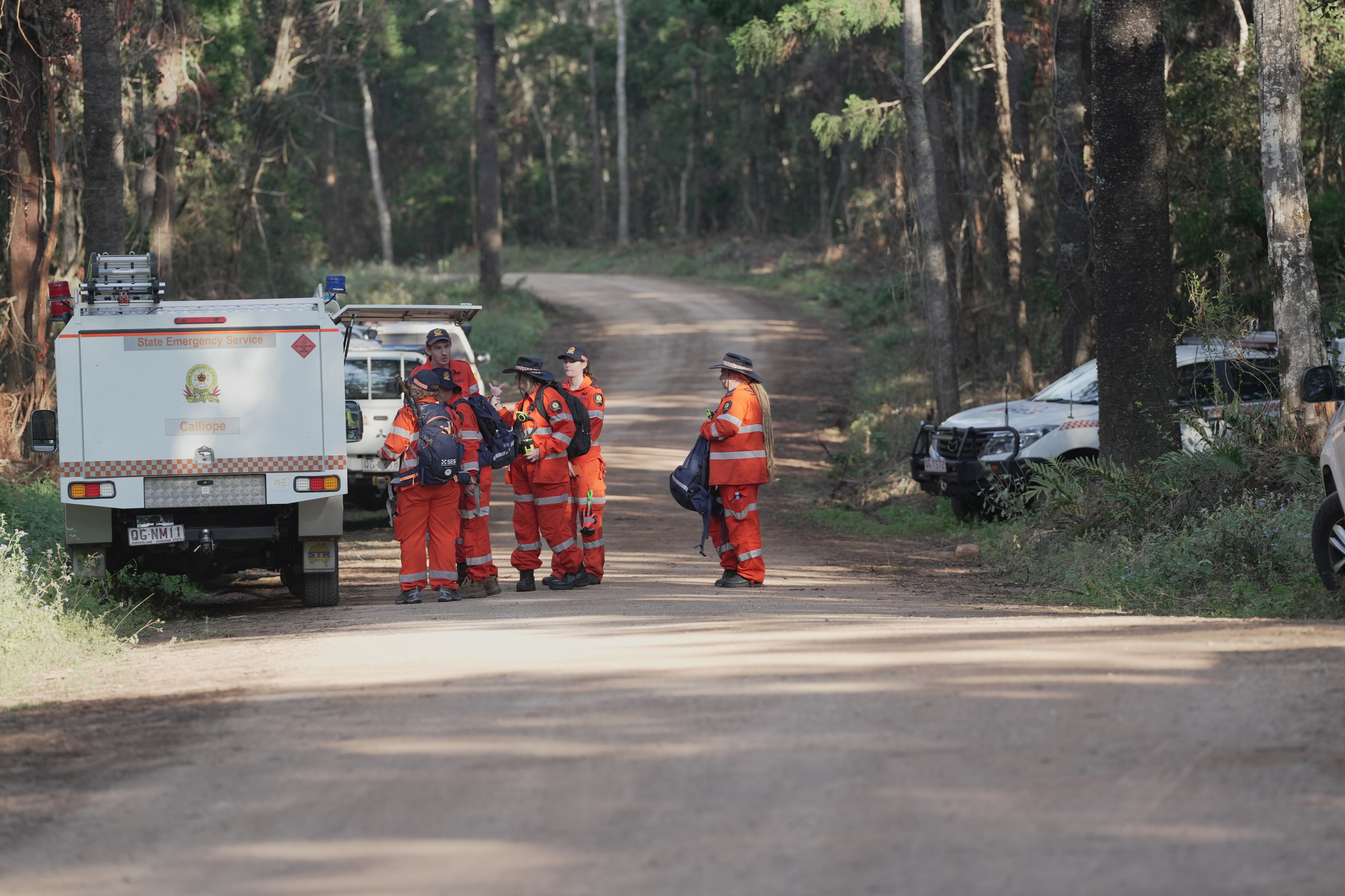 A group of people in orange jumpsuits by emergency vehicles on a dirt road in bushland