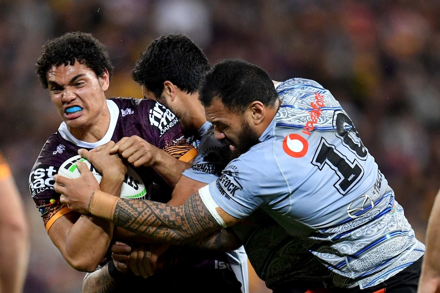 A male NRL player stands up in the tackle of two opposition players while he holds the ball.