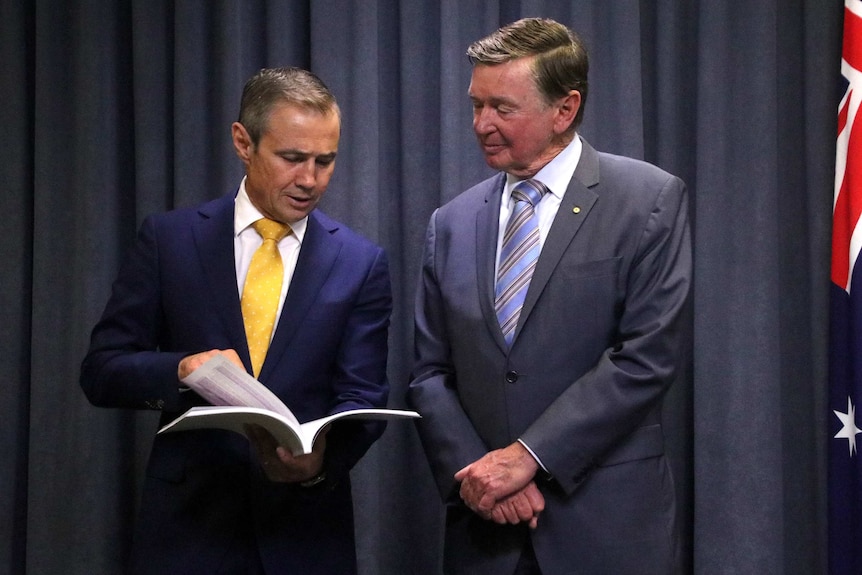 A mid shot of Roger Cook and Malcolm McCusker standing inside looking at a report dressed in suits.