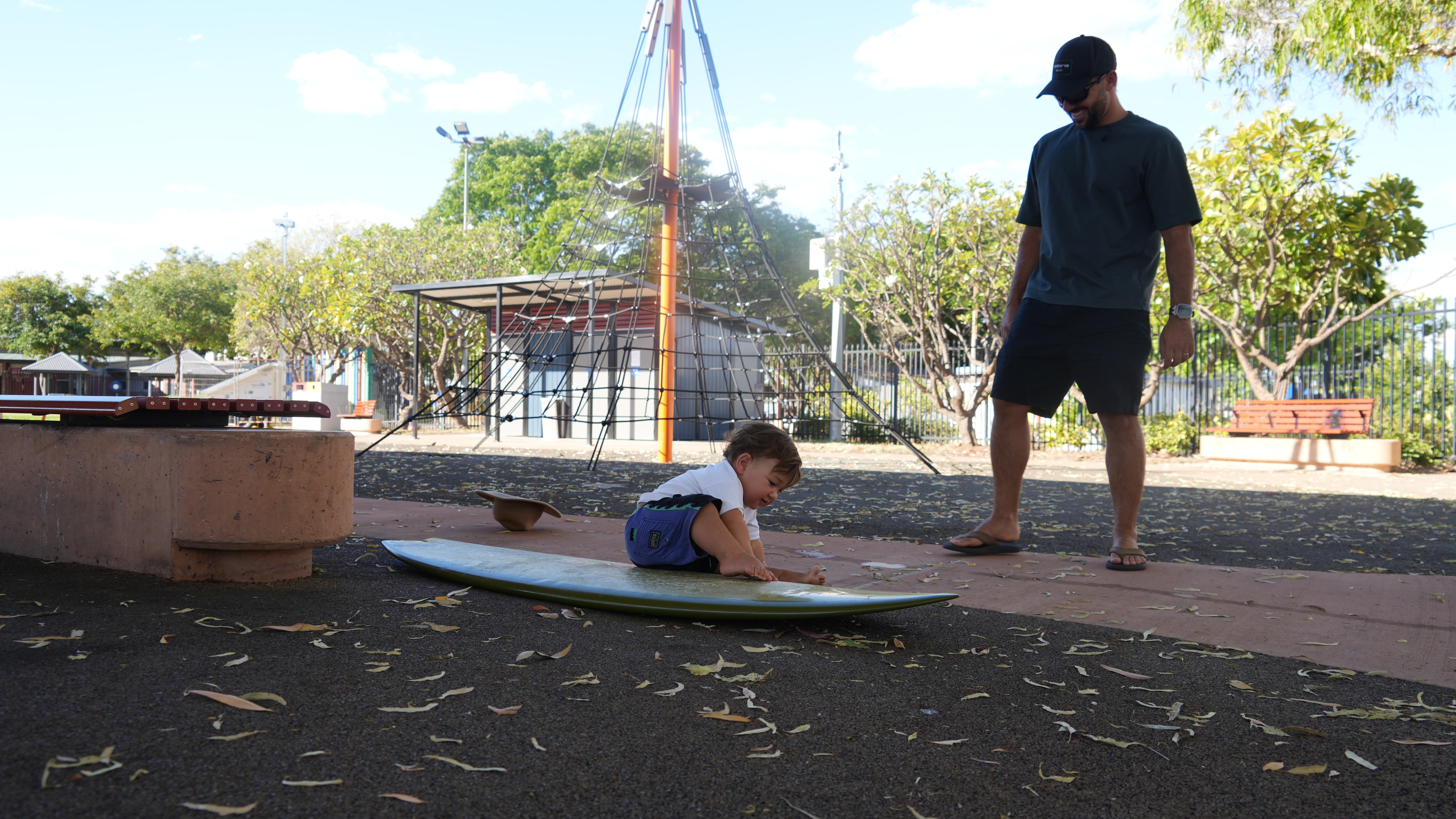 toddle sitting on surfboard with father watching behind, smiling