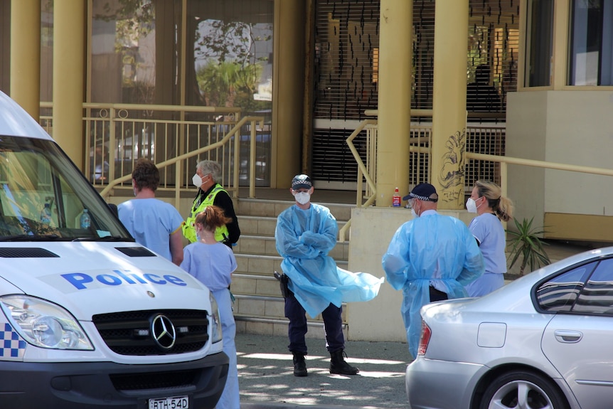 Emergency services dressed in personal protective equipment stand out the front of a building.